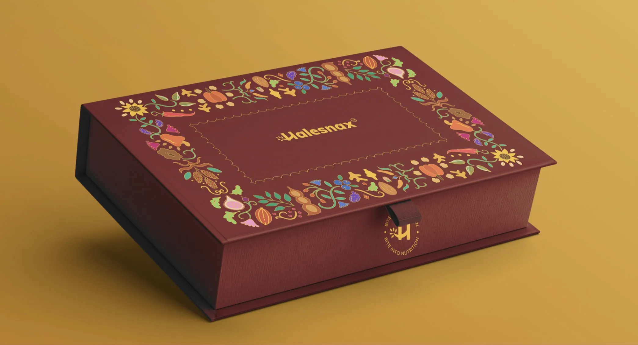 The gift box mockup on a mustard ground: a deep maroon book-style box, its lid carrying the border of gold-edged illustrated ingredients and the Halesnax logo, with a ribbon tab and a gold seal on the front.