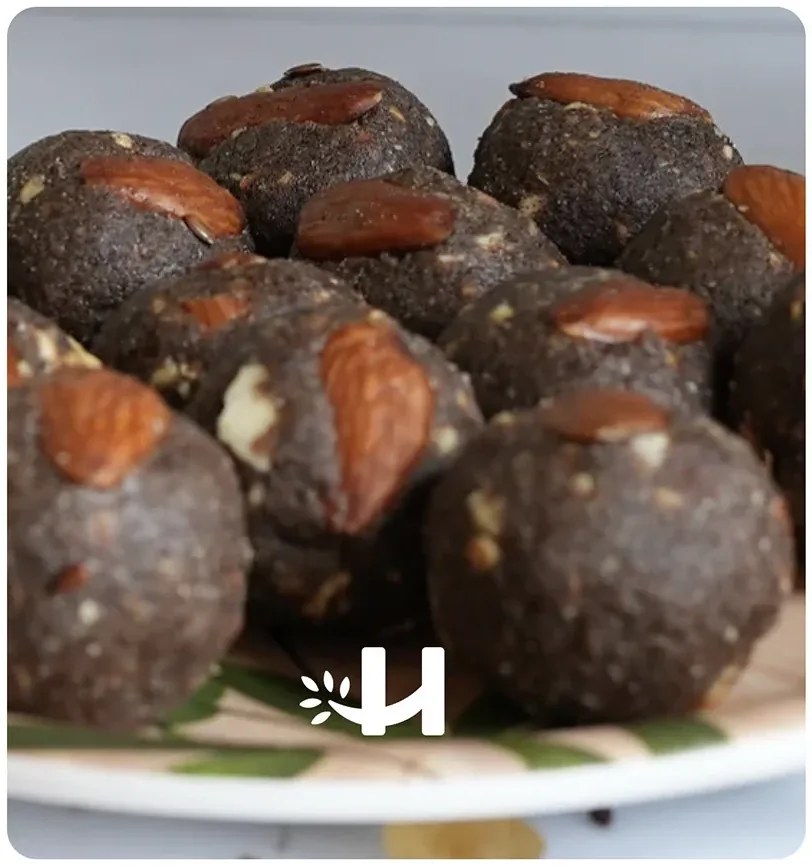 A close-up of dark laddoos topped with whole almonds on a leaf-printed plate, with the white H mark in the corner
