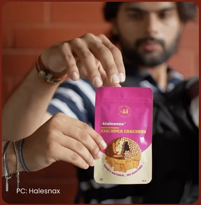 A bearded man holding up the pink Baked Chickpea Crackers pouch between his fingers, with the credit PC: Halesnax in the corner