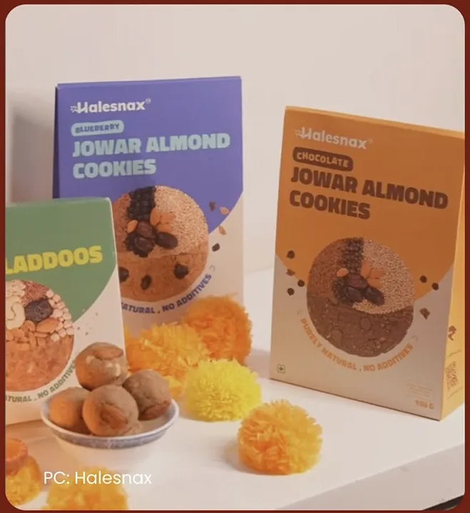 Blueberry and chocolate cookie packs and a laddoo box on a white counter, with marigolds and laddoos in front and the credit PC: Halesnax in the corner