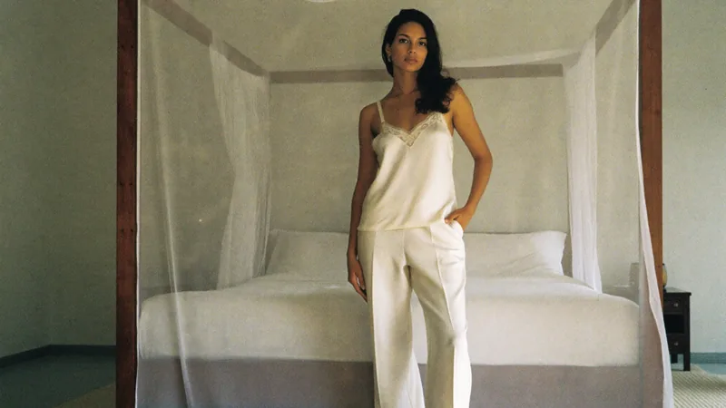 The shade seeker, in a satin camisole and trousers before a white canopy bed, morning